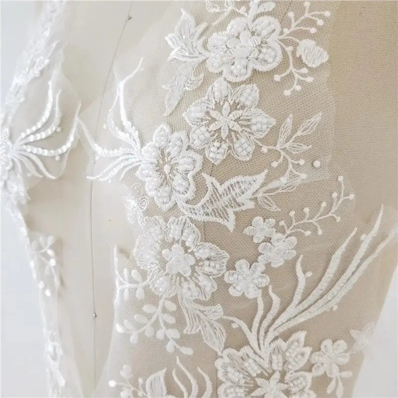 Robe de mariée blanche en dentelle avec application assortie, parée d’un tissu de dentelle florale blanche présentant des fleurs brodées délicates et des motifs de feuilles, rehaussés de petites perles de type perlé.