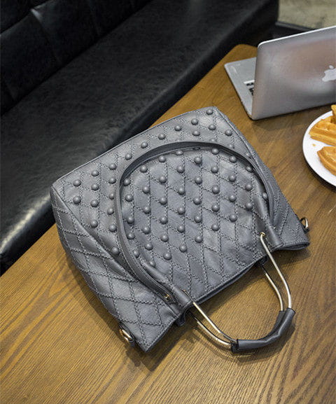 Bolso de hombro de cuero acolchado gris con tachuelas plateadas, asas de metal y un compartimento interior para un almacenamiento organizado.