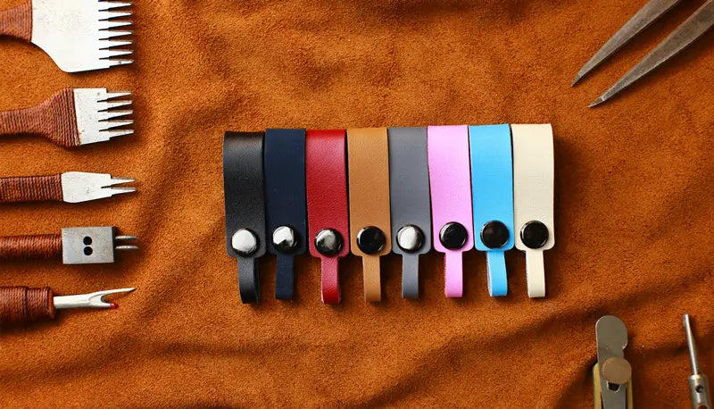 A row of seven genuine leather car keychains with metal key rings featuring snap button closures in black navy blue red tan gray pink light blue and cream colors.