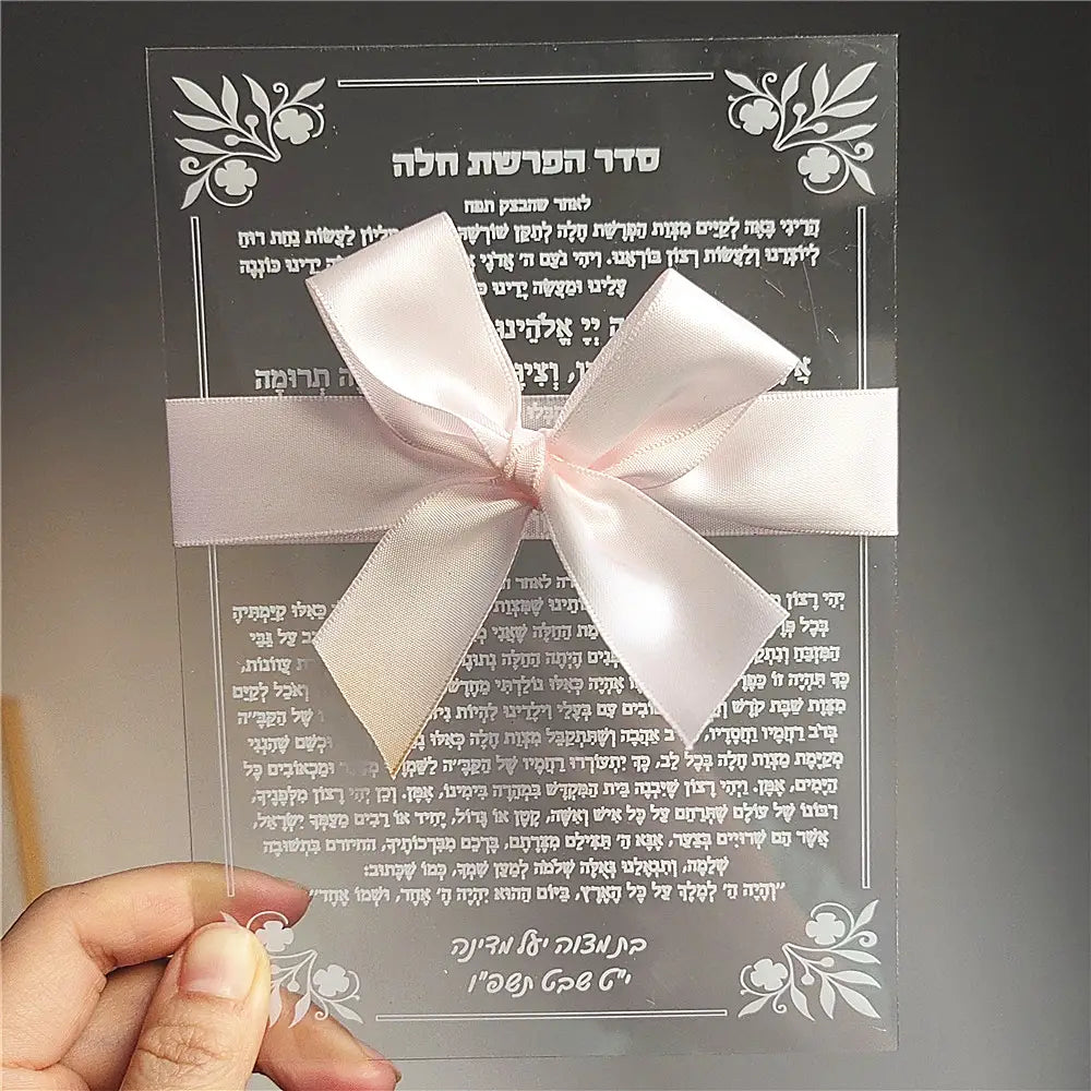 A delicate pink satin bow tied neatly across a transparent acrylic custom Hebrew prayer card with engraved text and floral designs for wedding and Bar Mitzvah souvenirs.