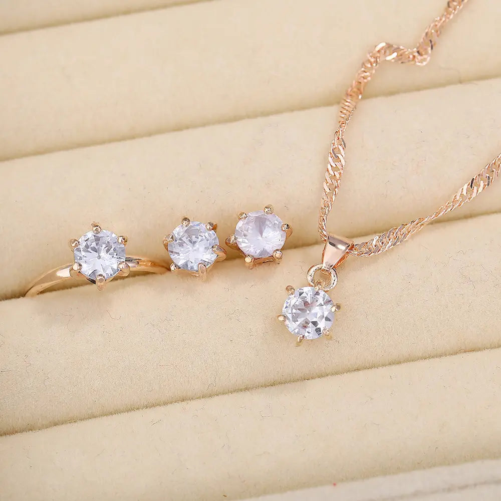 Juego de joyería de 5 piezas con circonitas cúbicas de color dorado rosado, que presenta un anillo, aretes tipo botón y un collar con dije, cada uno adornado con piedras de circonita cúbica transparentes.