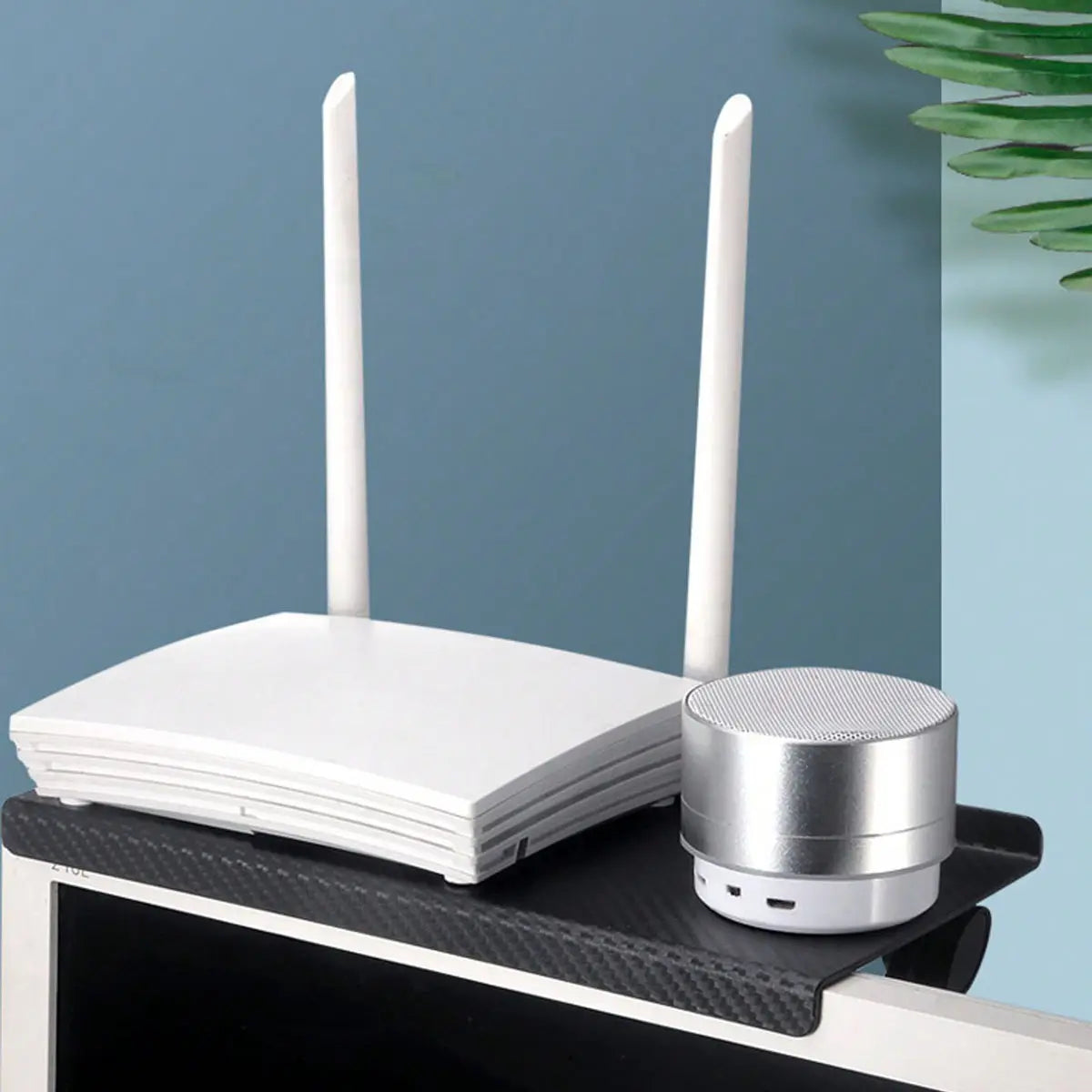 white wireless router with two vertical antennas and a silver cylindrical bluetooth speaker placed on a black carbon-fiber textured desk mat beside an adjustable screen top storage shelf monitor stand