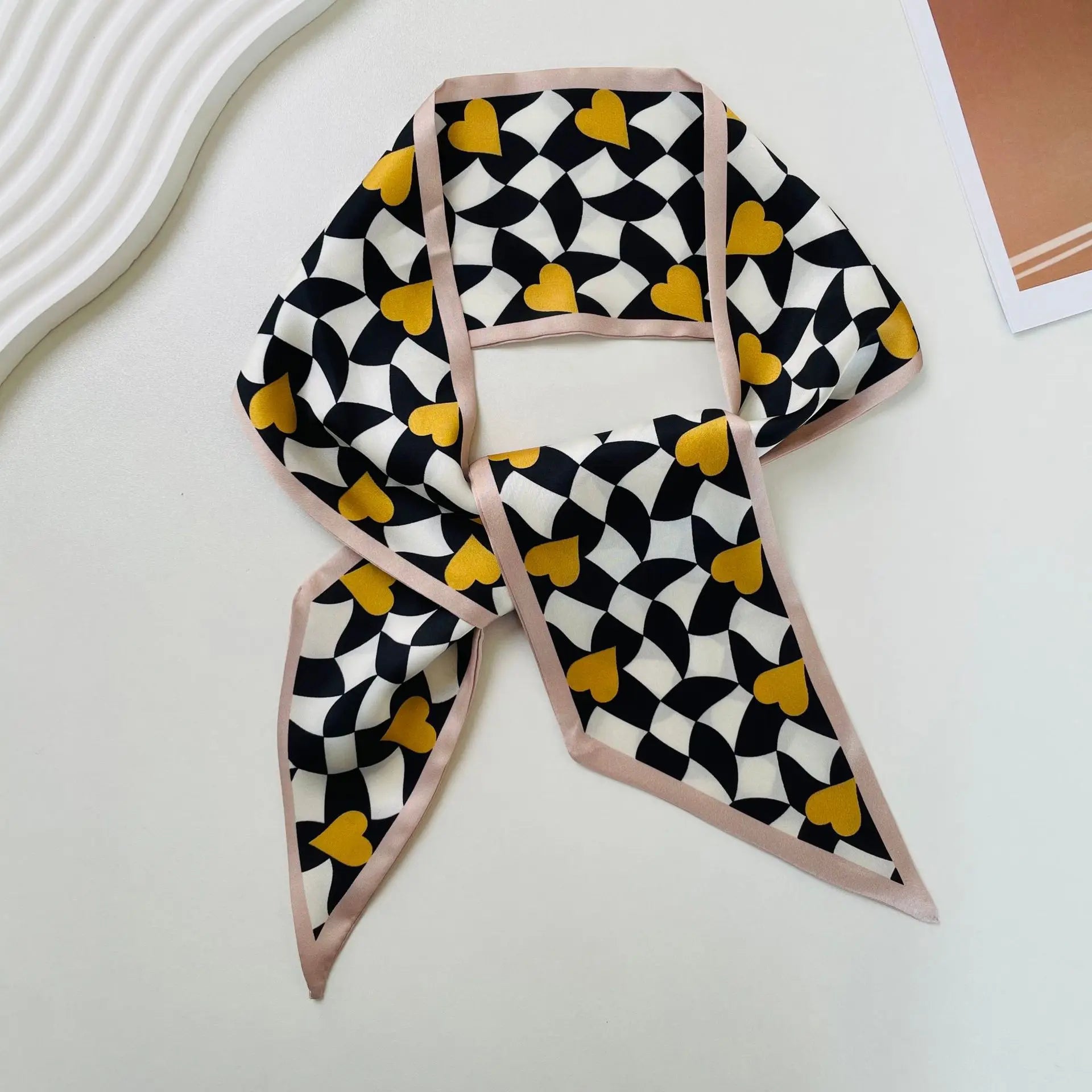 Un pañuelo de seda elegante en negro, blanco y amarillo mostaza con patrón de corazones, con un borde rosado melocotón suave, diseñado como una banda de escote cuadrado con estampado geométrico.