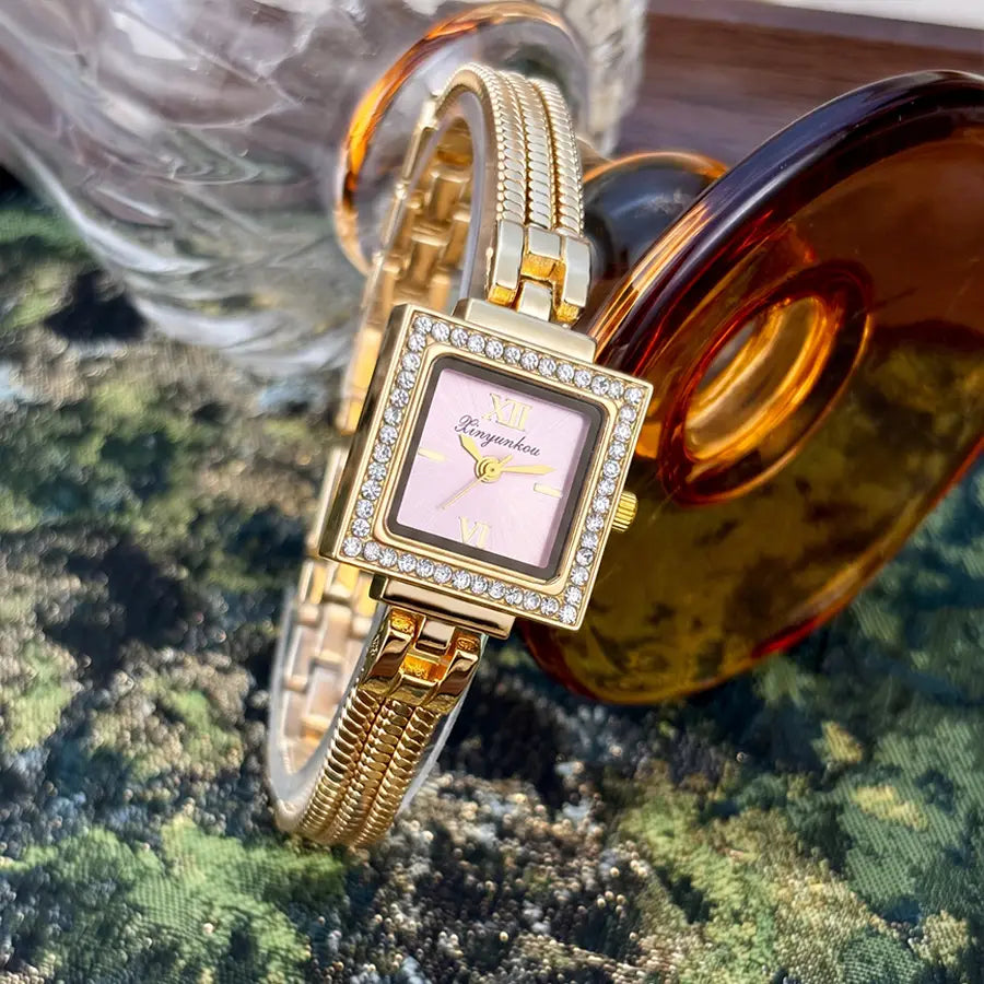 Gold diamond square watch with a gold-toned case, pink face, roman numerals, and diamond-studded bezel featuring a vintage fringe strap.