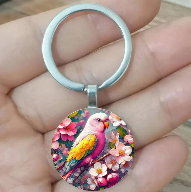 A lovebird keychain glass dome bird keyring with a vibrant pink and yellow parrot perched on a branch amidst blossoming flowers encased in a circular glass dome with a silver-toned metal ring.