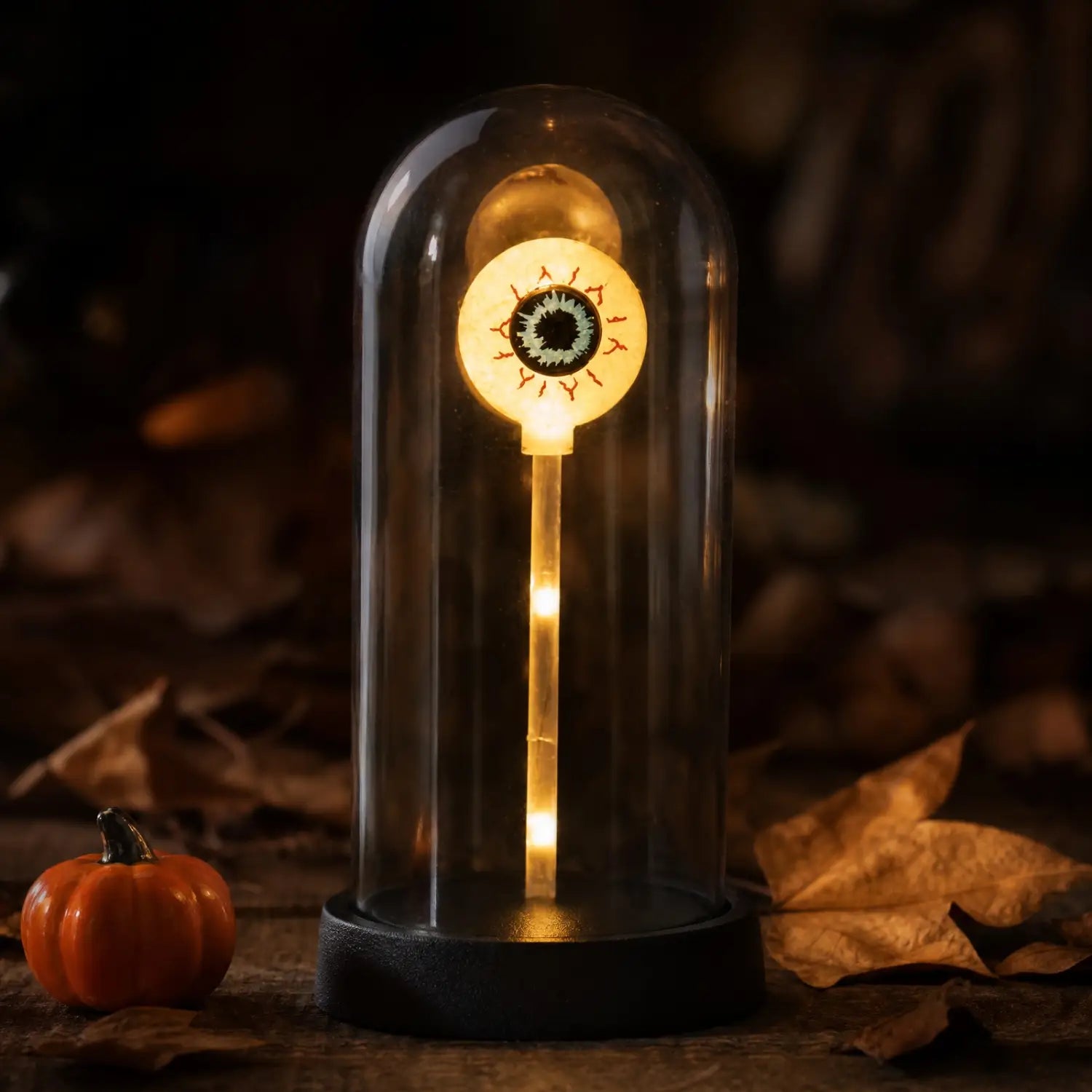 A glowing halloween-themed eye lamp made of glass with an illuminated stem and base encased in a clear dome featuring halloween lights decorations.