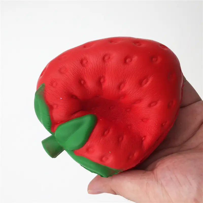 Un juguete antiestrés vibrante de fresa roja con textura, con forma de fresa, que se eleva lentamente y es blandamente esponjoso, con una parte superior de hojas verdes, sostenido en una mano.