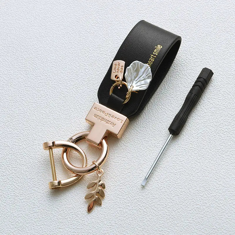 Llavero de piel negra con acentos metálicos de tono dorado rosado y colgantes en forma de hoja, diseñado como un llavero antirrobo estilo francés para automóvil.