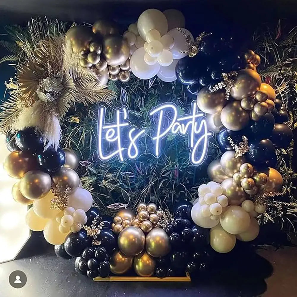 Ein schwarzes und goldenes Ballonbogen-Set für Partys mit einem leuchtenden Neon-„Let’s“-Party-Schild in geschwungener Schrift; mit schwarzen, goldenen und cremefarbenen Ballons sowie dekorativem Grünzeug.