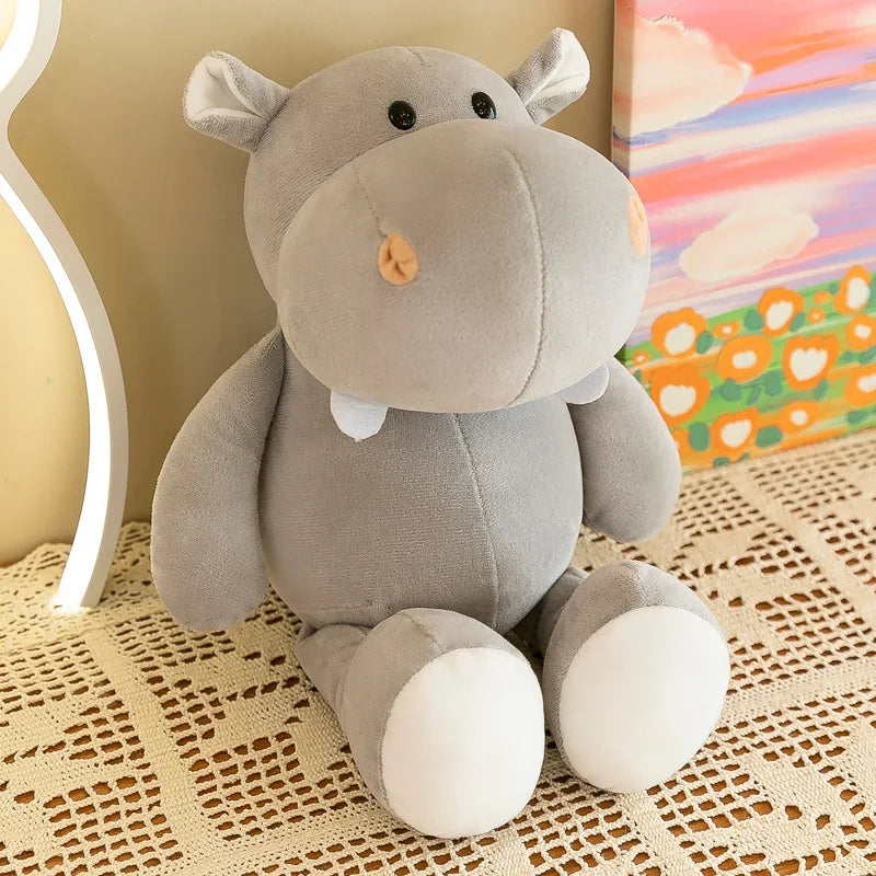 Un suave peluche de hipopótamo gris claro con pies blancos y una pequeña nariz tipo botón naranja, de la colección de animales de peluche de la jungla.