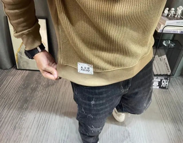 Pull-over en maille à empiècement contrastant pour homme, manches longues, en tissu de maille côtelée beige, avec une étiquette blanche « kuh radio » sur l’ourlet.