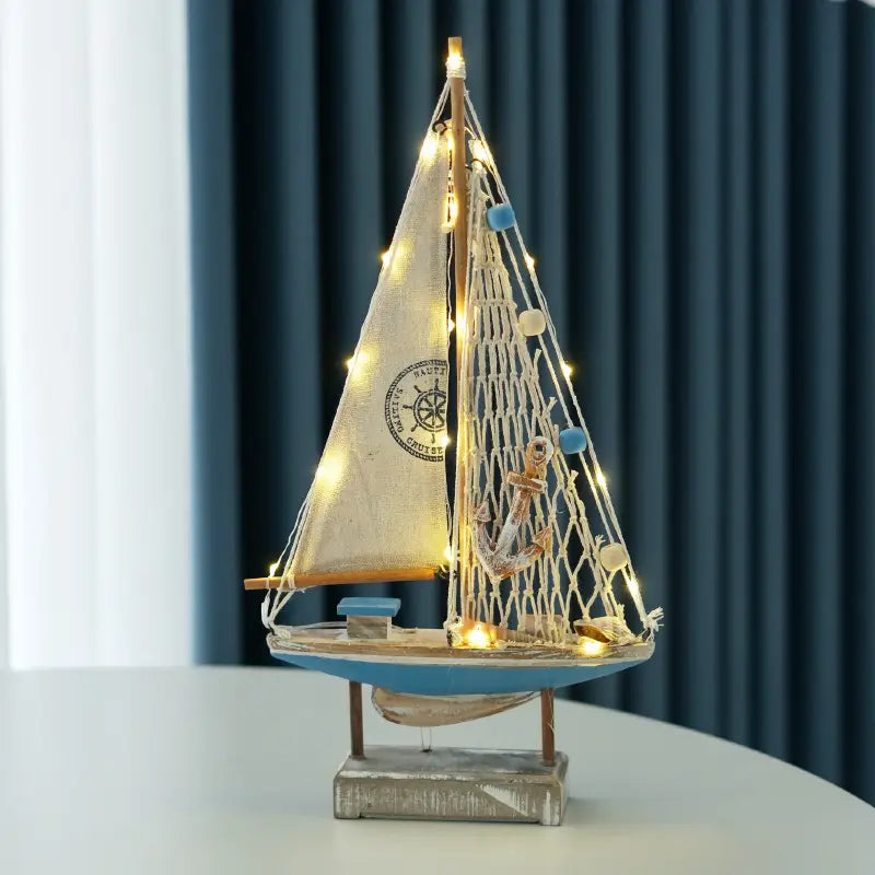 Deko im mediterranen Stil mit einer hölzernen LED-Segelboot-Dekoration für das Zuhause: ein dekoratives maritimes Modell-Segelboot mit beiger Segelleinwand, blauem Rumpf und hölzernem Mast, geschmückt mit Lichterketten sowie kleinen blauen und weißen Perlen.