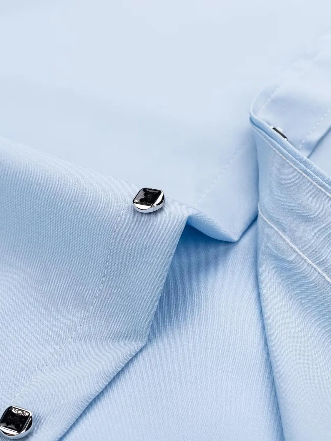 Silberfarbener eckiger Knopf mit schwarzer Einlage auf einem hellblauen Herren-Hemd mit langen Ärmeln im Business-Casual-Look aus elastischem, einfarbigem Stoff.