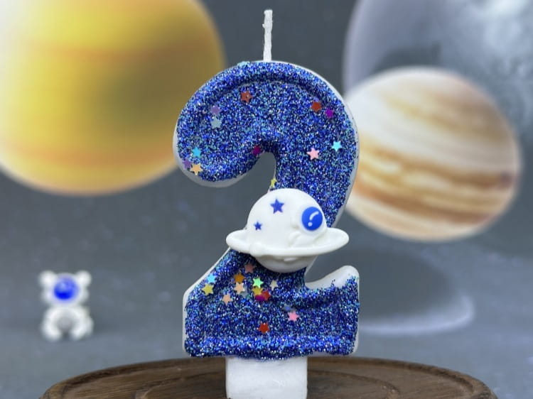 Una vela azul con brillo con el número dos para cumpleaños, con una figura de astronauta blanca y chispas de estrellas de colores, diseñada como adorno para pastel de fiesta planetaria.
