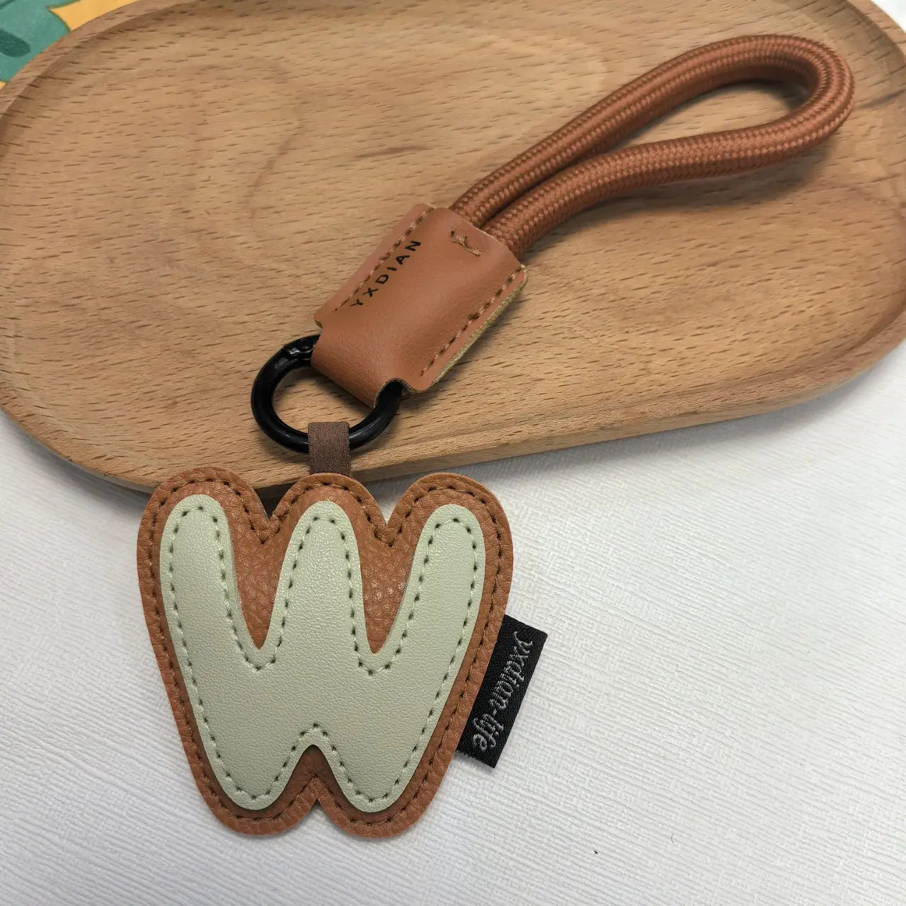 A brown and cream-colored leather initial keychain shaped like the letter W with stitched detailing and wristband charm.
