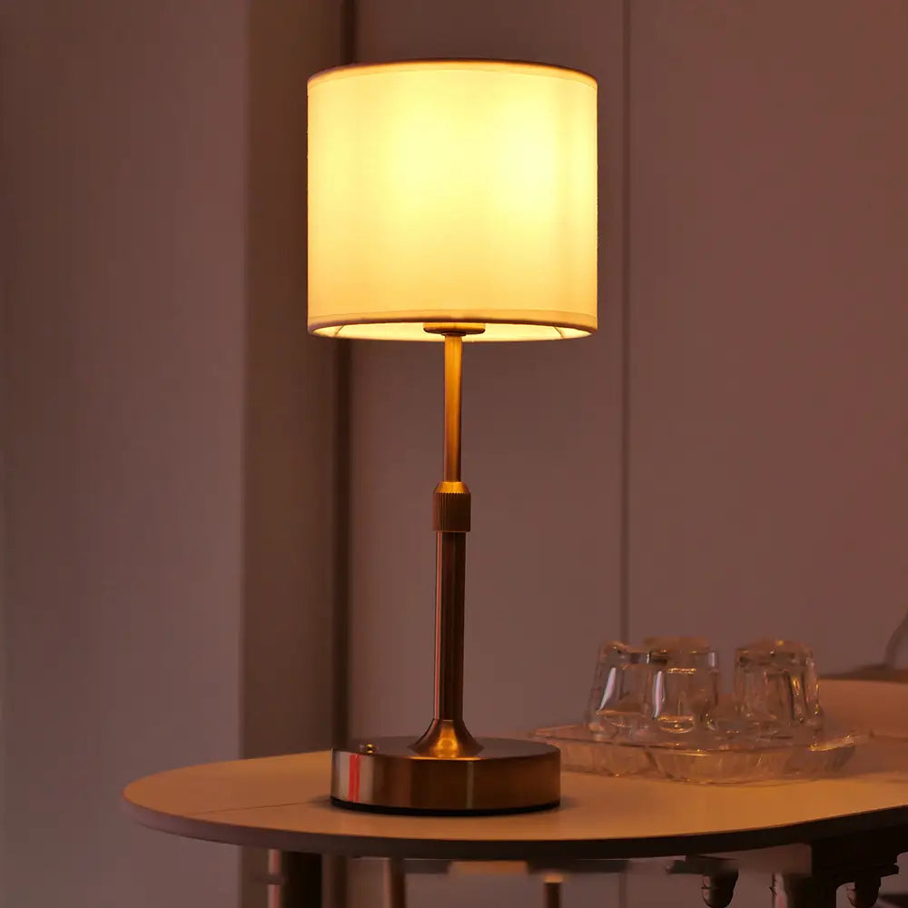 Warmtonige Touch-Tischlampe mit einer messingveredelten Metallbasis und -stange sowie einem weichen cremefarbenen Stoffschirm, von vorn dargestellt.
