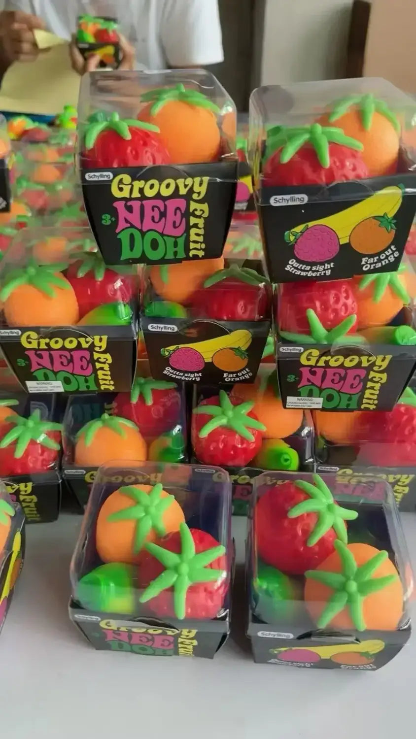 Eine Frontansicht von grell bunten, groovigen NEE-DOH-Obst-förmigen, quetschbaren Spielzeugen mit orangefarbenen Tomaten und roten Erdbeeren mit grünen Blättern in klarer Kunststoffverpackung aus dem Erdbeere-Orange-Banane-Stressabbau-Set.