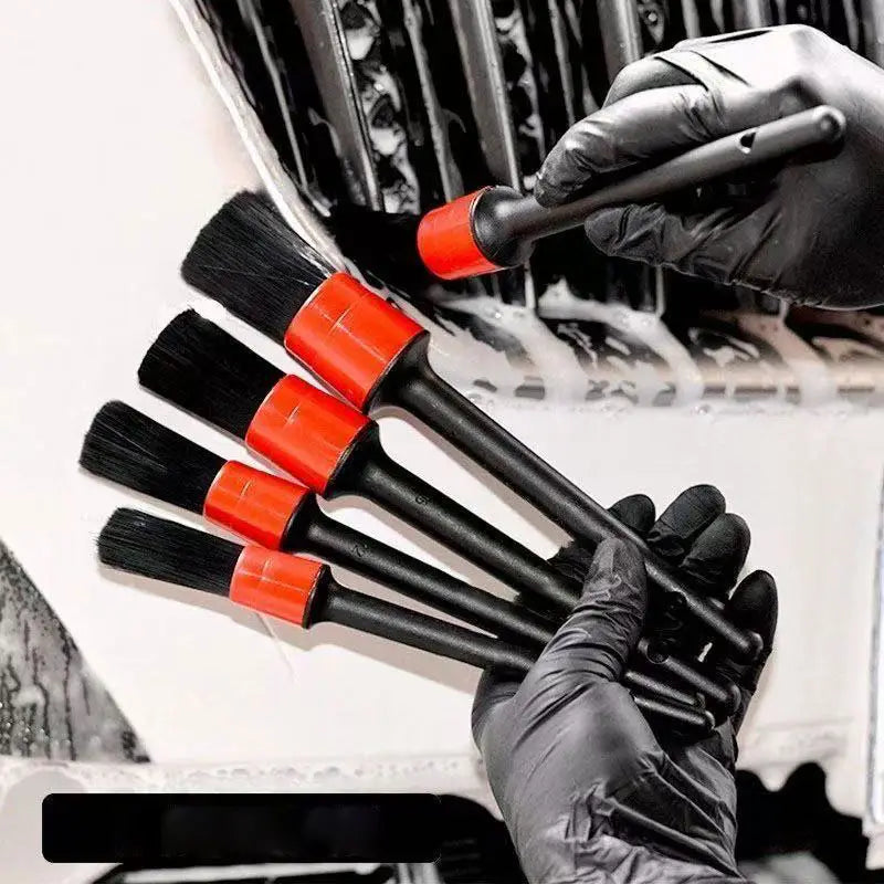 Ensemble de cinq brosses de nettoyage pour voiture, avec des soies noires et des manches en plastique rouge éclatant, tenues dans des mains gantées contre un arrière-plan de véhicule savonneux, pour le nettoyage et la finition.