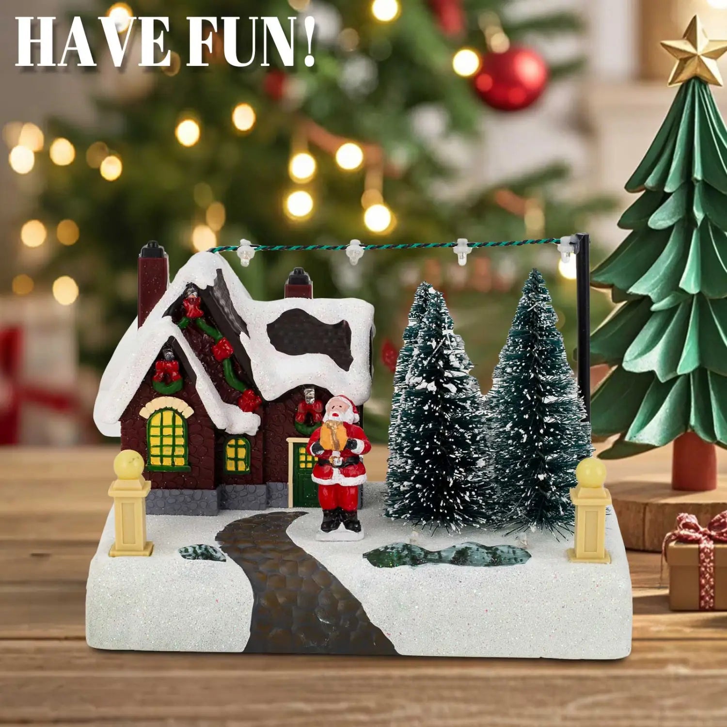 サンタと雪を備えたLEDクリスマスビレッジの家。緑の窓のある赤と白の雪化粧をしたミニチュアの家、霜のかかった松の木が2本、雪の小道にクラシックな装いのサンタクロースの置物が配置された様子。