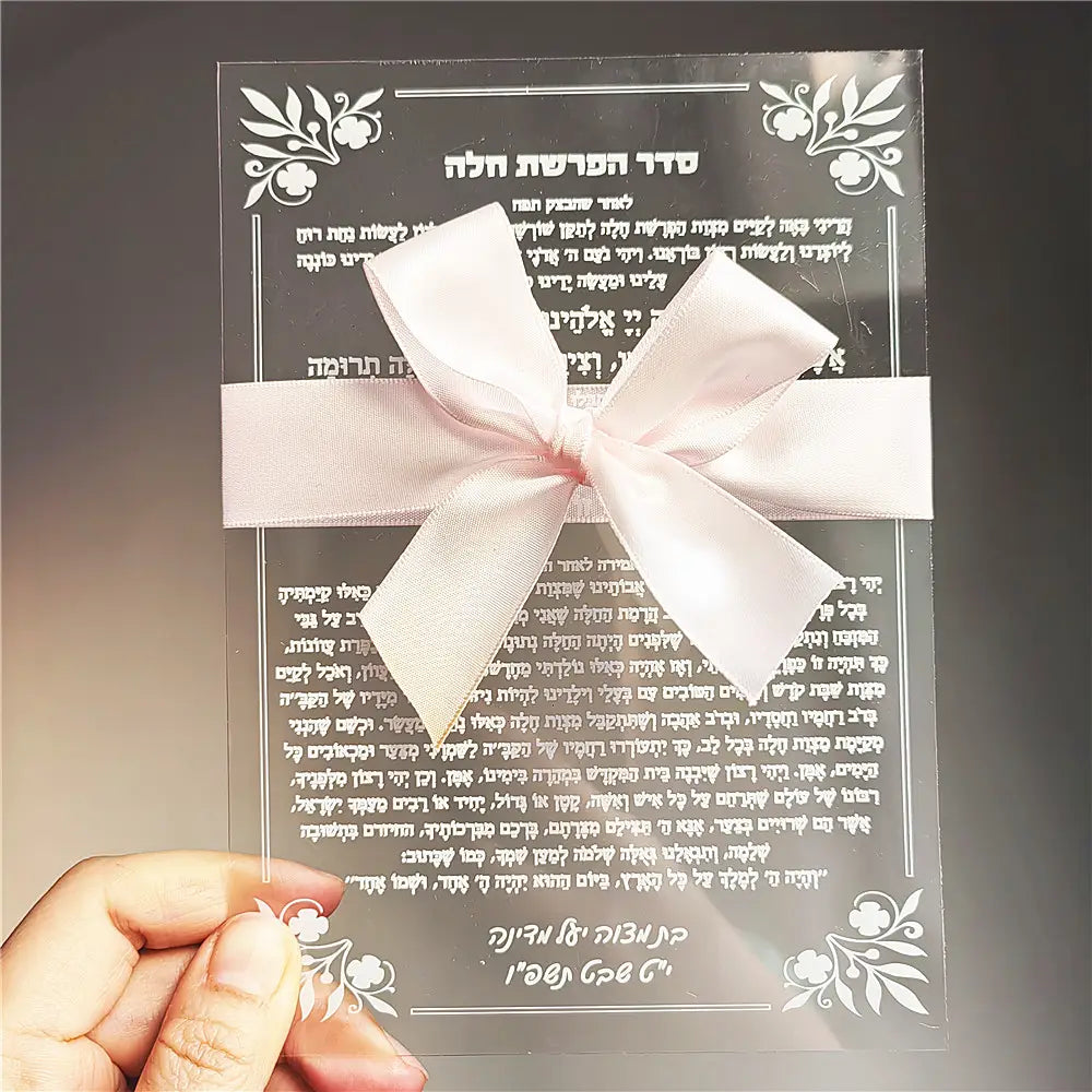 A delicate white satin bow tied neatly across a transparent acrylic card with engraved Hebrew text and floral designs for a custom Hebrew acrylic prayer card wedding and Bar Mitzvah souvenir.