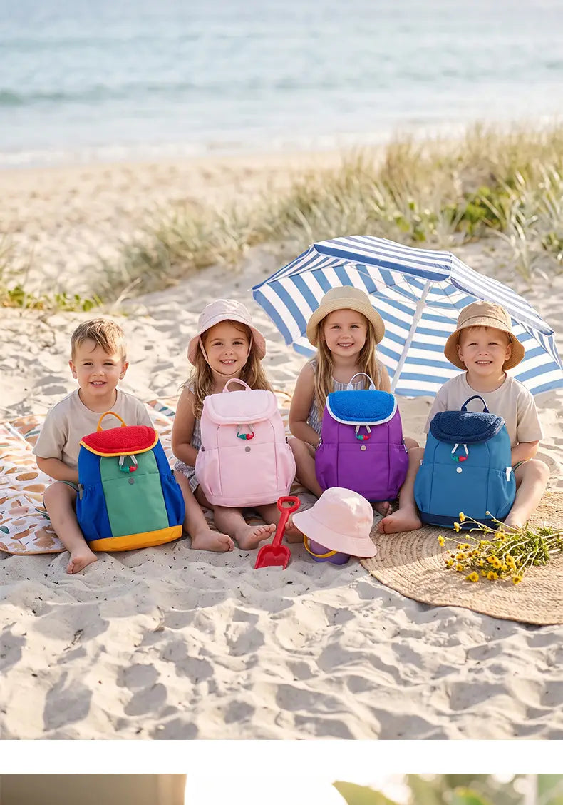 Vier Kinder sitzen am sandigen Strand, tragen Hüte und sitzen neben bunten leichten Fuzzy-Rucksäcken für Kinder; entworfen als Kindergartentagesrucksäcke mit verspielten Designs.