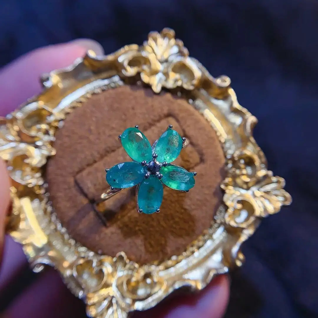 Ein zarter Smaragdblumen-Silberring mit einem feinen silberfarbenen Ring, der fünf leuchtende, smaragdgeschliffene grüne Edelsteine in einer Blumenform trägt, ergänzt durch einen kleinen dunkelblauen Akzentstein in der Mitte.