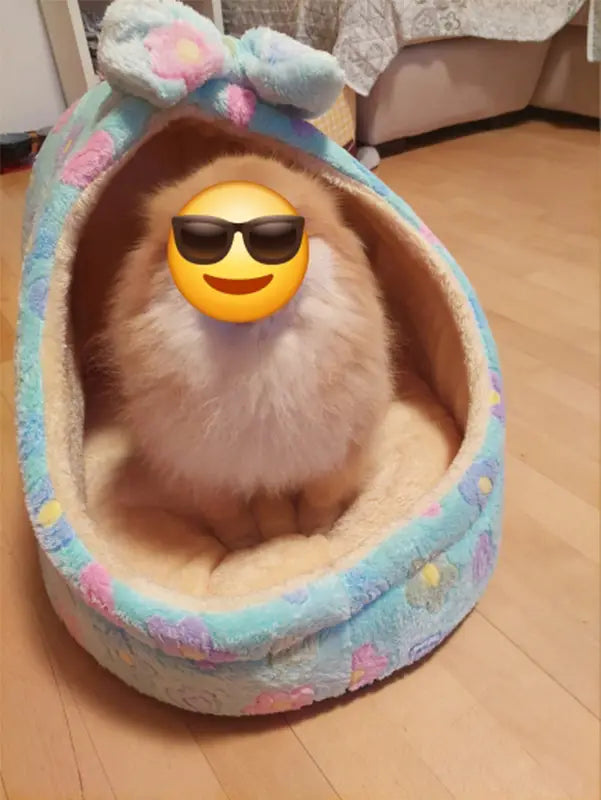 A fluffy light-colored dog sits comfortably inside a foldable plush pet cave bed for small dogs and cats featuring a cozy pastel blue and pink floral pattern with a soft bow on top.