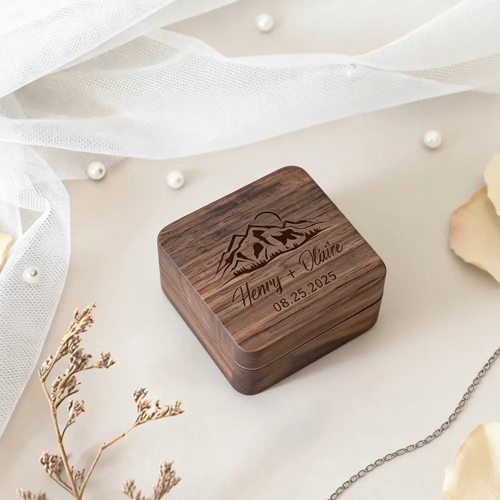 A personalized wooden wedding ring box with a natural wood grain finish engraved with a mountain design and the names Henry plus Claire and the date 08.25.2025.