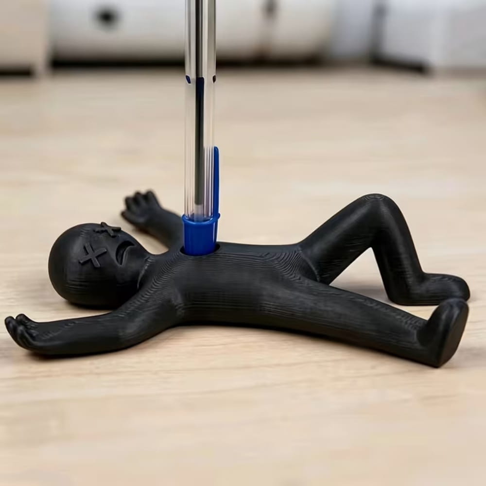Black plastic stick figure pen holder desk organizer shaped like a reclining figure with x’s for eyes holding a blue ballpoint pen.