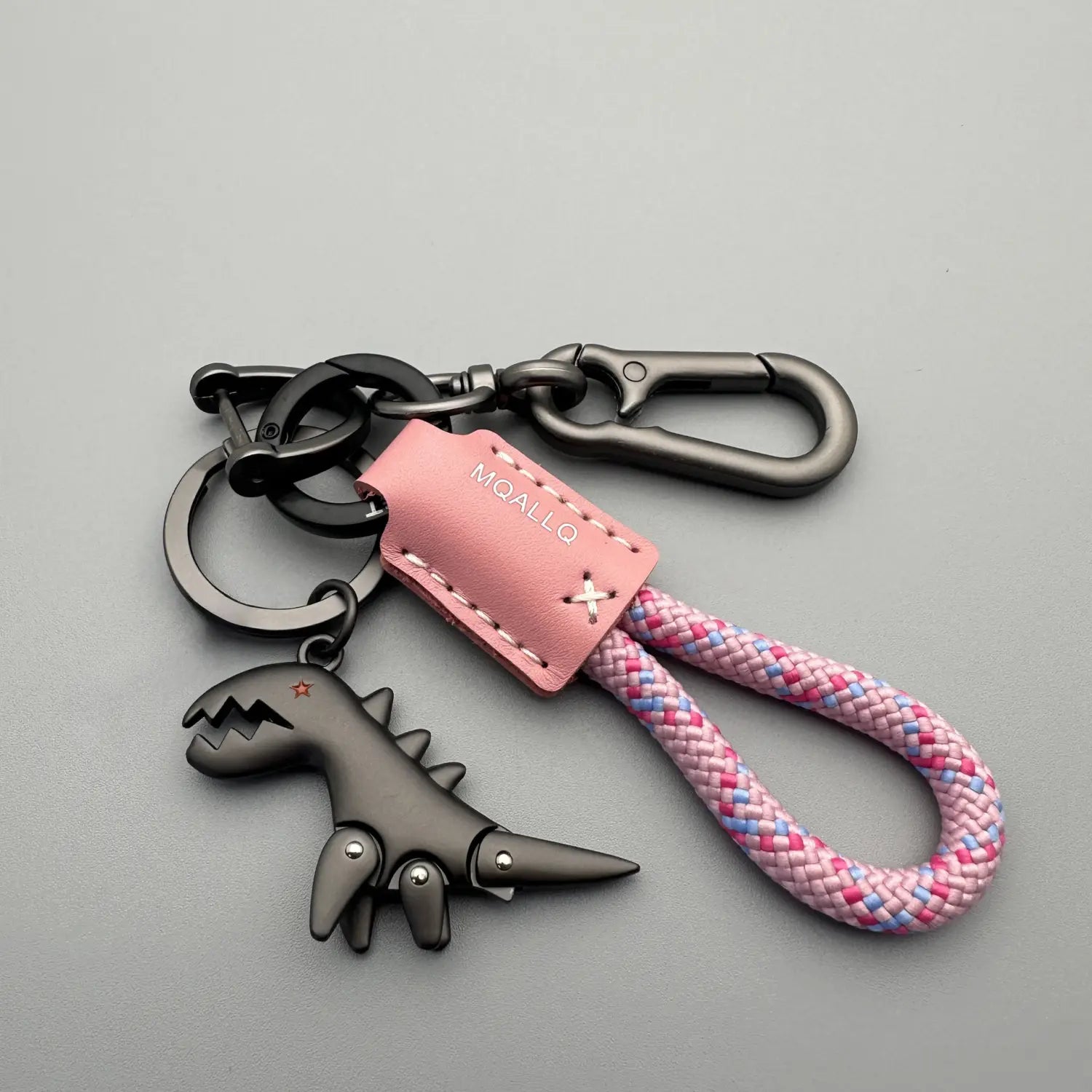 llavero de cuero con forma de dinosaurio trenzado en tonos rosa y azul, llavero metálico, con un adorno de dinosaurio negro que presenta remaches plateados y un broche mosquetón metálico de acabado mate.
