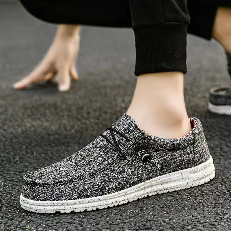 Chaussures décontractées de type mocassins à enfiler en toile pour hommes, en toile texturée grise, avec des lacets noirs et une semelle en caoutchouc blanche à motifs d’aspérités.