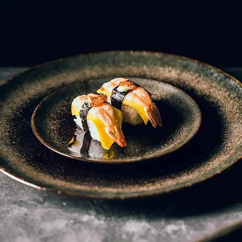 Two pieces of shrimp nigiri sushi topped with yellow fish and crispy orange flakes on a rustic Japanese ceramic dinner plate suitable for traditional tableware use.