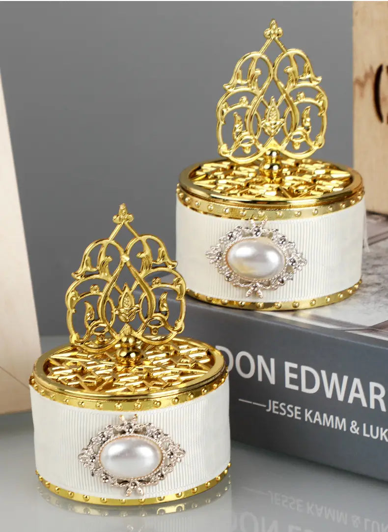 Boîte à bonbons de mariage dorée décorative élégante blanche cannelée, avec un couvercle bordé d’or, un dessus en filigrane doré orné et une perle centrale entourée de strass étincelants, conçue comme contenant de perle-bonbon décorative.