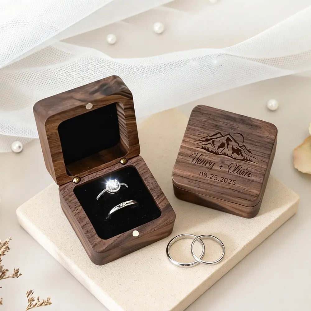 Personalized dark wood engraved wedding ring box with mountain and date design holding a diamond engagement ring and wedding band.