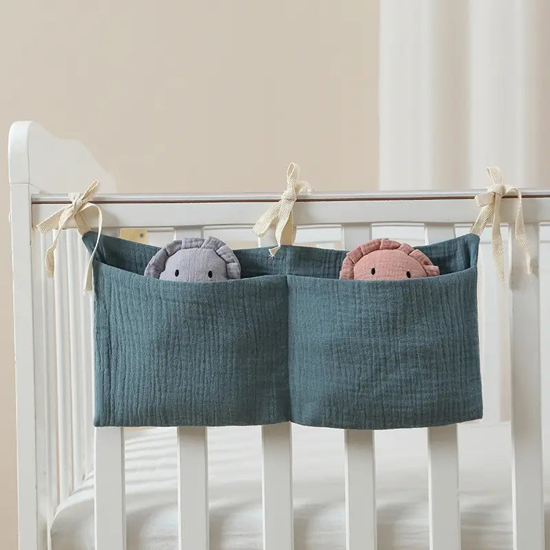 Tei­alfarbene Aufbewahrungstasche für Babybettchen aus Baumwollstoff mit zwei Fächern, mit gebundenen Schleifenbändern aus Stoff, hält zwei plüschgefüllte Kuscheltiere als Organizer neben dem Bett.
