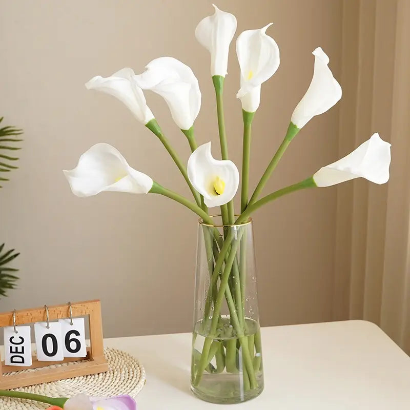 A bouquet of white calla lily flowers with green stems arranged in a clear glass vase for home decor.