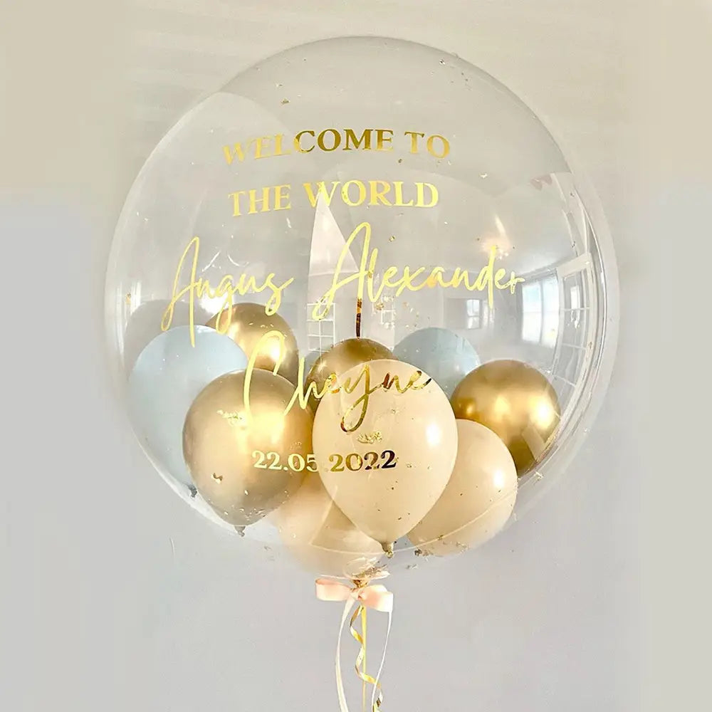 Individueller Baby-Party-Ballonaufkleber mit goldenem Text auf einem klaren transparenten 36-Zoll-Bobo-Ballon, gefüllt mit gold- und cremefarbenen Ballons, personalisiert mit den Namen August Alexander Cheyne sowie dem Datum 22.05.2022.