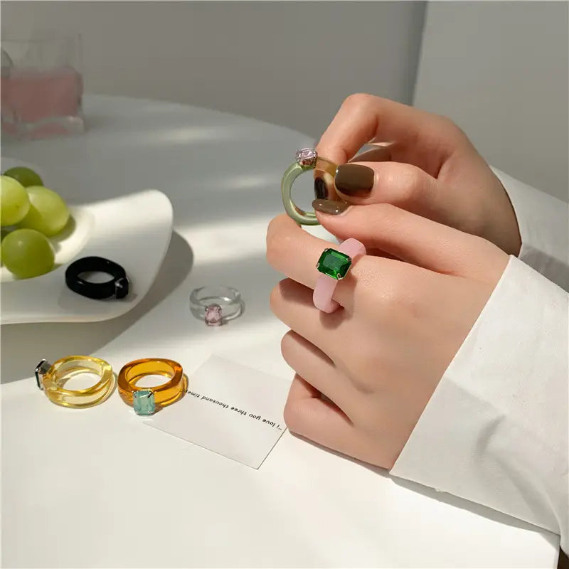 A hand holds a colorful resin crystal geometric ring for women featuring a pastel pink resin band with a square-cut green gemstone in its center.