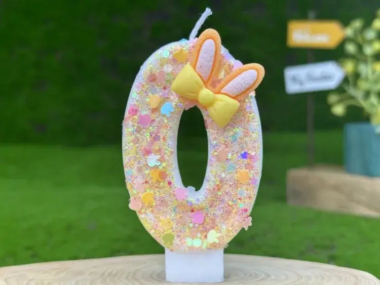 A glitter bunny ear number birthday candle featuring a white glittery zero adorned with a yellow bow and orange bunny ears.