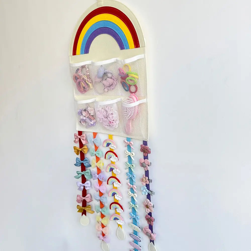 Organizador colgante para accesorios de cabello con diseño temático de arcoíris, bolsillos de malla y cintas adornadas con lazos colgantes, montado en la pared.