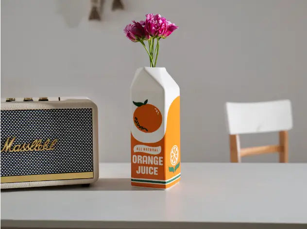 Un vase en céramique blanc et orange, façonné comme une brique de jus, avec une étiquette imprimée comportant une illustration d’orange, des accents verts et le texte « all natural orange juice ».
