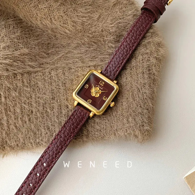 A square women quartz watch with a gold-tone case, burgundy textured leather strap, and maroon dial featuring golden hour markers and a floral design at the center.
