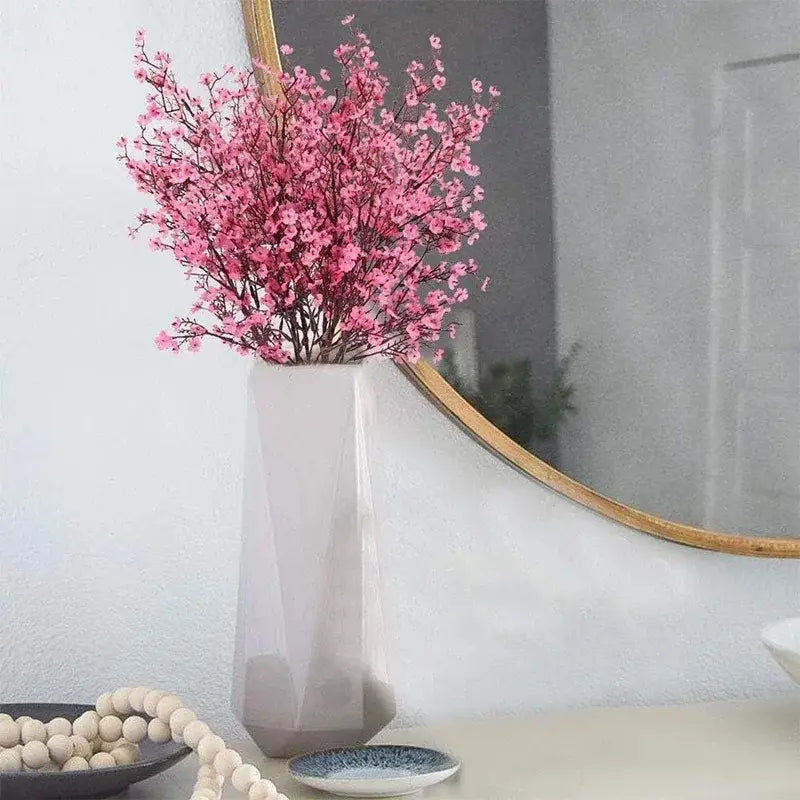 A sleek white ceramic vase with geometric facets holding a vibrant bouquet of pink artificial flowers cherry blossoms.
