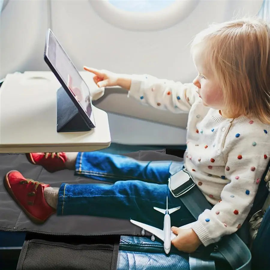 Ein Kind in einem weißen Pullover mit Punkten und blauen Jeans sitzt auf einem Flugzeugsitz und nutzt eine Kinder-Flugzeug-Fußstütze-Reisebett-Sitzverlängerung; auf der ausklappbaren Ablagetischplatte hält es ein Tablet.