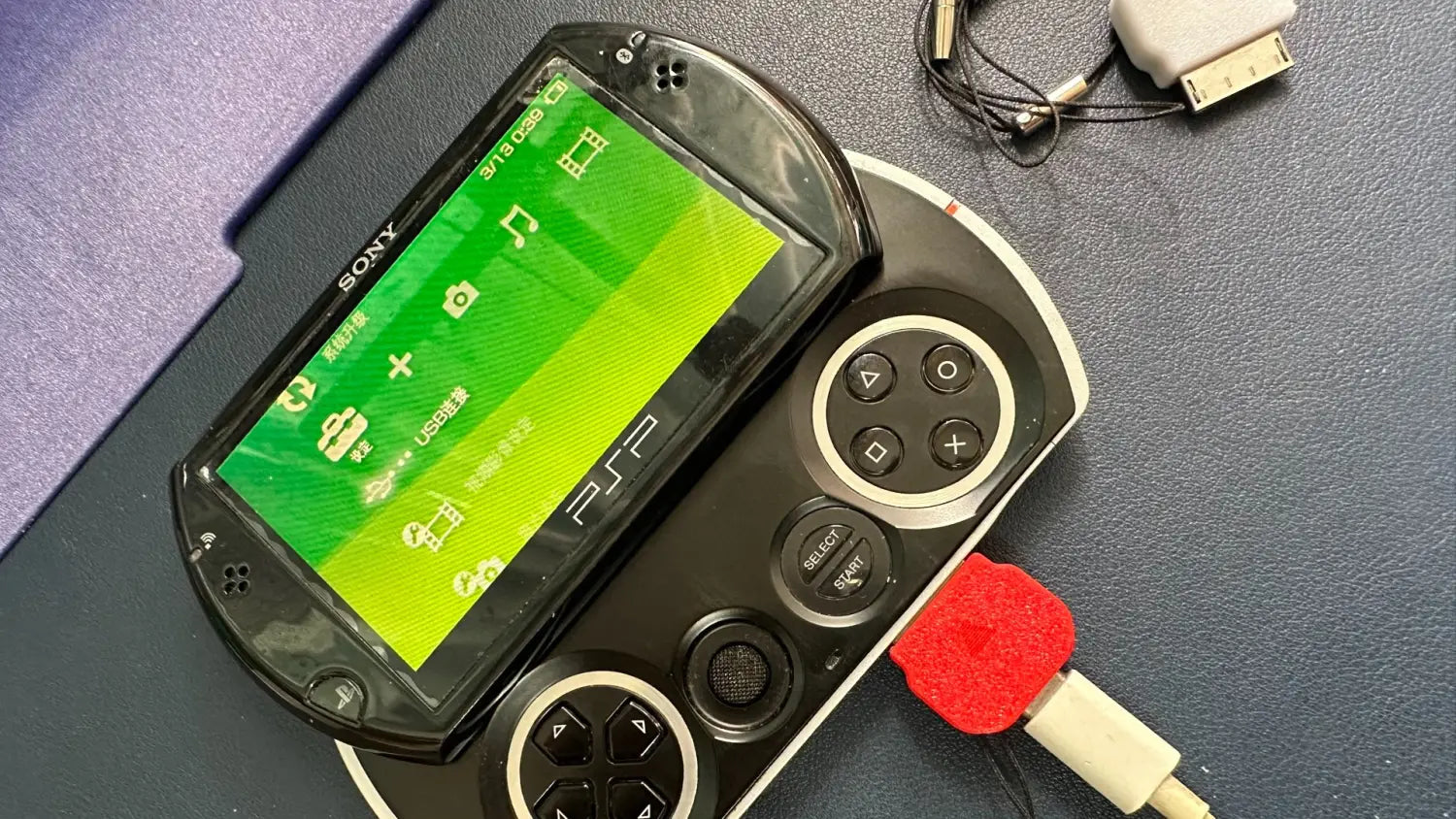 デュアルアナログスティックおよび方向ボタンを備えたブラックのSony PSP Go携帯型ゲーム機。緑色の画面にアイコンとPS2ロゴを表示。PSP GoタイプCコンバーター充電ケーブル用アクセサリーを備えています。
