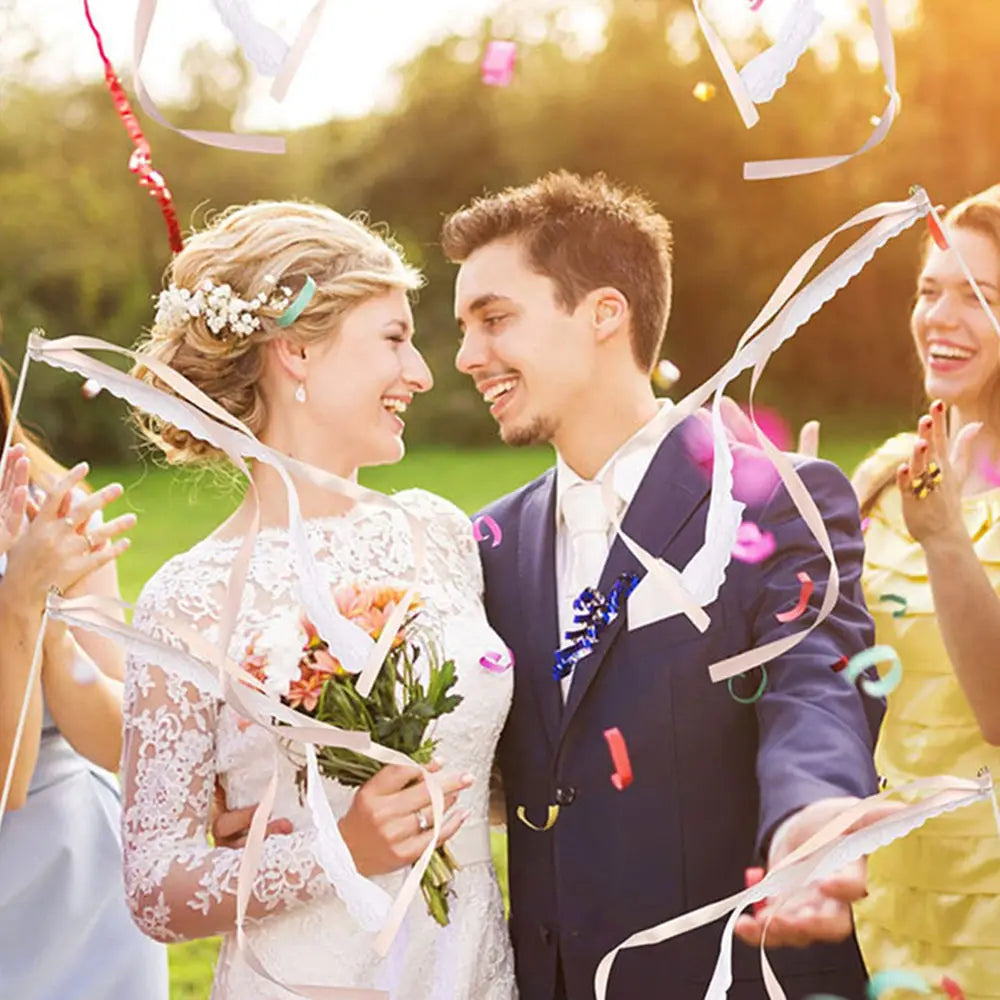 La mariée porte une robe de mariée en dentelle blanche à manches longues et à motif floral raffiné, à côté de baguettes de ruban de mariage avec dentelle et cloches.