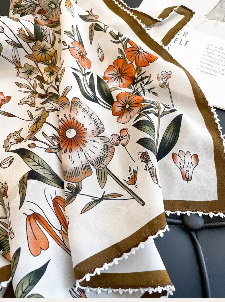 A delicate cream-colored silk floral square scarf featuring vibrant orange blossoms green leaves and brown accents bordered by gold trim and finished with a soft white lace edge.