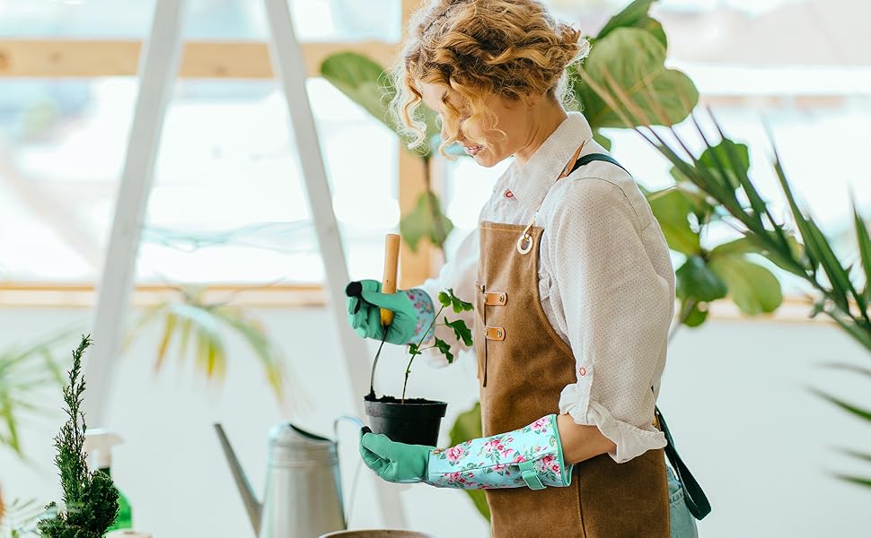 Une femme portant un tablier en cuir marron tient une petite plante en pot tout en portant des gants de jardinage floraux marron respirants pour femmes, anti-épines.