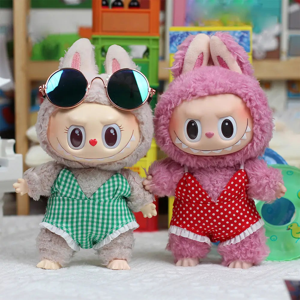 Dos figuras de muñeco Labubu de peluche del conjunto de bañador de verano para muñeco Labubu de 17 cm, con un atuendo gris esponjoso con pantalones cortos de cuadros vichy verdes y gafas de sol redondas, y un pijamita esponjoso rosa con pantalones cortos de lunares rojos.