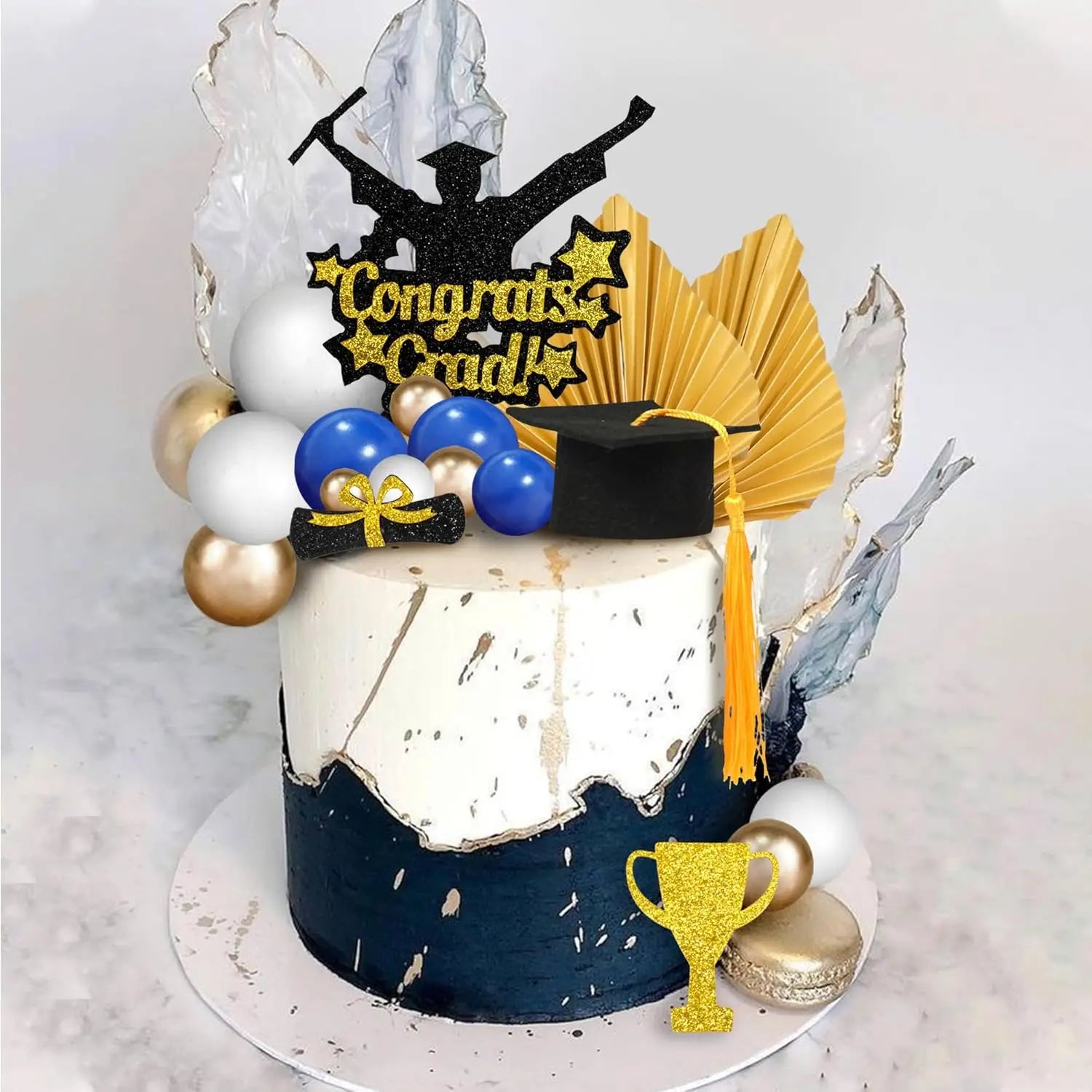 a graduation cake topper set for 2026 senior party featuring a white and navy blue speckled cake design topped with glittery gold stars, blue white and gold balloons, a black graduation cap, and a congrats grad topper